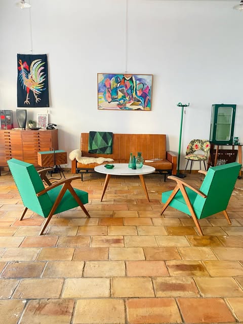 Un salon vintage avec deux fauteuils verts, un canapé en teck, une table basse et des tableaux colorés sur tomettes